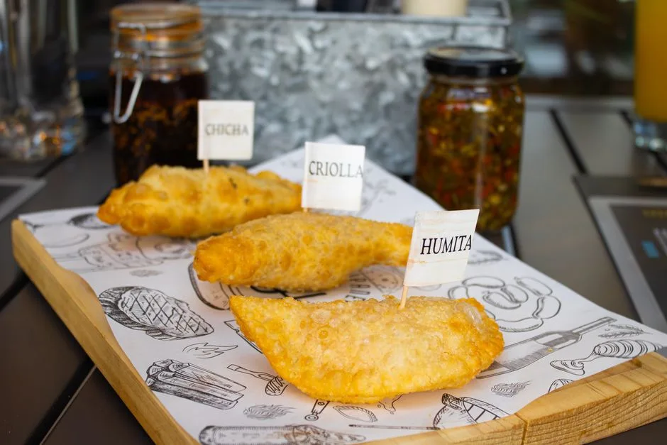 Un trío de empanadas etiquetadas, servidas en una bandeja, que muestran sabores tradicionales como chicha, criolla y humita.